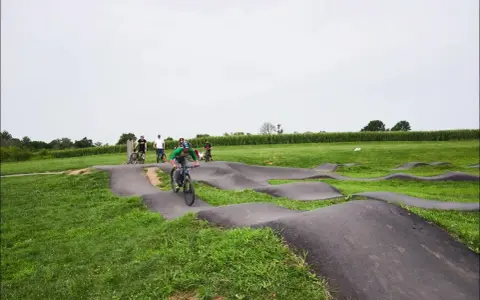A group of people enjoy riding bikes on a winding pump track, with rolling hills and a cornfield in the background.