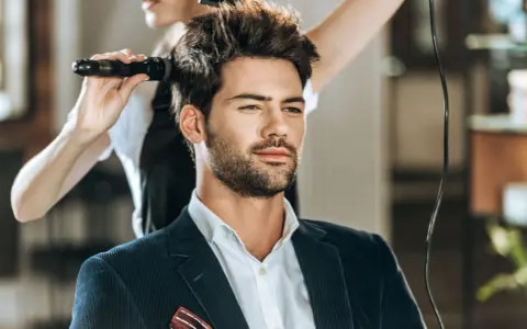 Mężczyzna w eleganckim garniturze czeka na stylizację fryzury męskiej. Fryzjerka suszy jego włosy suszarką.