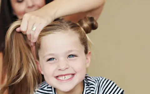 Ein Mädchen mit blauen Augen und Sommersprossen lächelt, während ihre Mutter ihr die Haare zu niedlichen Dutts frisiert. Tolle Ideen für kinder frisuren!