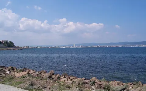Słoneczny brzeg jak pić. Widok na morze i miasto w oddali, z kamienistym wybrzeżem na pierwszym planie.