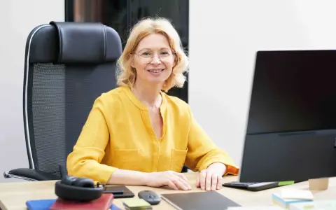Uśmiechnięta wicedyrektor w okularach siedzi przy biurku, przed nią komputer i słuchawki.