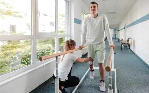 Młody mężczyzna ćwiczy chód przy poręczy, a fizjoterapeutka pomaga mu w rehabilitacji.