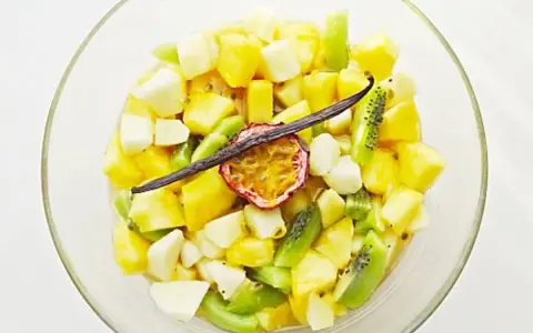 Salade de fruits tropicaux coupés en dés dans un bol en verre, garnie de fruits de la passion et d'une gousse de vanille.
