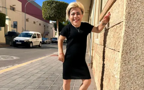 Mujer sonriente con vestido negro, apoyada en pared de piedra. Sus medidas cuerpo mujer son únicas y hermosas.