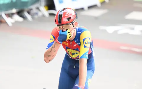 Radfahrer Thibau Nys wischt sich mit der Hand über das Gesicht, trägt einen roten Helm und ein blau-gelbes Trikot.