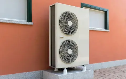 Unità esterna di una pompa di calore, con due ventole, montata su una base rialzata contro un muro arancione con finestre.