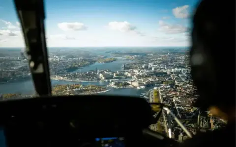 Vy från en helikopter över Stockholm. Staden breder ut sig vid vattnet under en klarblå himmel med moln.