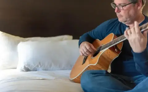 Mężczyzna w okularach gra na gitarze siedząc na łóżku. Gdzie ta keja nuty?