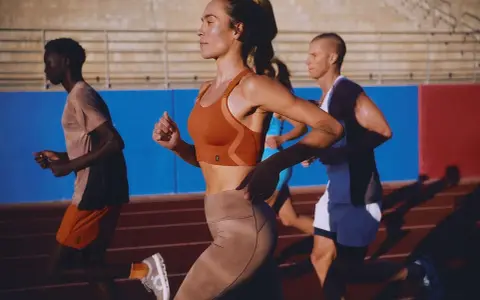 Biegacze na bieżni. Kobieta w pomarańczowym topie i beżowych legginsach, mężczyzna w koszulce i szortach. Buty sportowe z niskim dropem zapewniają komfort.
