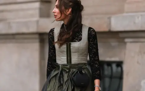 Eine Frau trägt eine graue Weste über einer schwarzen Spitzenbluse. Sie kombiniert die schwarze Bluse mit einem langen, olivgrünen Rock und einer kleinen schwarzen Tasche.