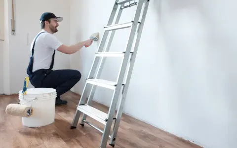 Hombre con mono azul y gorra usa brocha para limpiar paredes, junto a escalera y cubo con rodillo.
