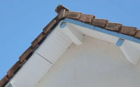 Le coin d'une toiture en pente avec des tuiles anciennes et un acrotère blanc.