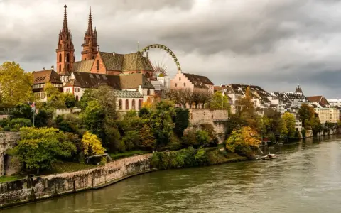 A scenic view of Basel's historic Minster and riverside buildings, perfect for a Basel itinerary. A Ferris wheel peeks out from behind the architecture.