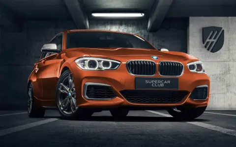 Pomarańczowe BMW 140i z tabliczką "SUPERCAR CLUB" stoi w garażu.