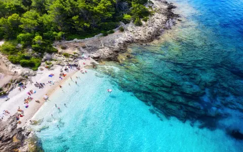 Idealne miejsce, gdzie lecieć w czerwcu: kamienista plaża z turkusową wodą, sosnowy las i opalający się ludzie.