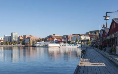 En vacker dag att besöka Luleå. En träspång leder längs vattnet förbi röda byggnader och en vit båt.