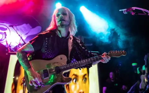 John 5 mit seinem charakteristischen Make-up spielt Gitarre, im Hintergrund ein Bild von Marilyn Manson.