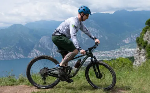 Mężczyzna w kasku jedzie na rowerze MTB po górskim szlaku. Widok na jezioro i miasto w oddali.