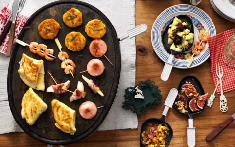 Raclette-Ideen für Kinder: Garnelen-Spieße, kleine Frikadellen, Käse-Speck-Röllchen und Crêpes auf einem Raclette-Grill.
