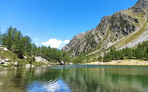 Il **lago d'Arpy** riflette il cielo azzurro e le montagne rocciose. Sulle rive, pini e persone godono del sole estivo.