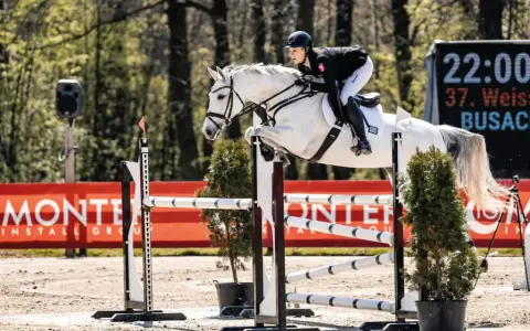 Greta Busacker und ihr weißes Pferd überwinden eine Hürde. Im Hintergrund eine digitale Anzeigetafel.