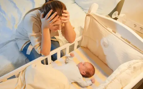 Madre agotada, con las manos en la cabeza, mira a su bebe que se desvela por la noche en su cuna.