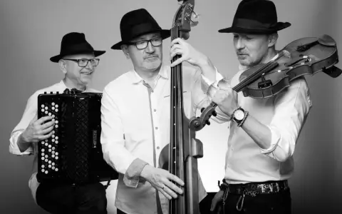 Trzech muzyków w czarnych kapeluszach grających muzykę klezmerską: akordeonista, basista i skrzypek.
