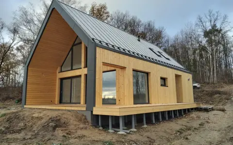Nowoczesny dom 70m2 z drewnianą elewacją i dużymi oknami, otoczony lasem.