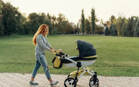 Ruda kobieta z uśmiechem pcha wózek podczas pierwszego spaceru z noworodkiem w parku.