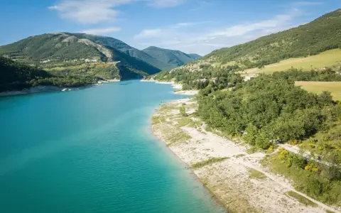 Splendido **lago di Fiastra** con acque turchesi, circondato da colline verdi e case sparse. Un paesaggio sereno e incantevole.