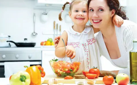 Mama i córka przygotowują sałatkę. W kuchni widać warzywa, oliwę i chleb. To zdrowa żywność, bogata w oligosacharydy, które wspierają florę bakteryjną.