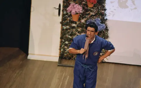 Actor con mono azul y pañuelo a cuadros, actuando en un teatro para niños. Fondo con flores y enredaderas.