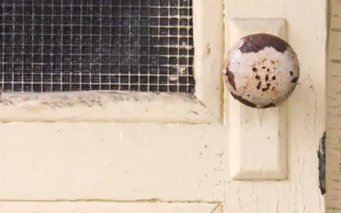 Maniglia usurata su porta interna tamburata, pronta per essere rinnovata. La rete della finestra è visibile.