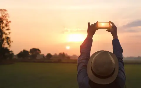 Osoba w kapeluszu fotografuje zachód słońca telefonem z aparatem szerokokątnym.