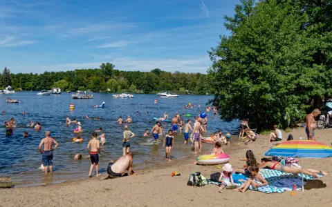 Menschen genießen das Reschensee baden an einem sonnigen Tag. Kinder planschen, Erwachsene schwimmen und entspannen am Ufer. Boote gleiten über das Wasser.