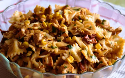 Pyszna sałatka z kurczakiem i makaronem farfalle, kukurydzą i suszonymi pomidorami, posypana natką pietruszki.
