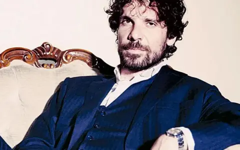 L'attore Montanari, con barba e capelli ricci, indossa un completo blu e una camicia bianca, seduto su una poltrona d'epoca.