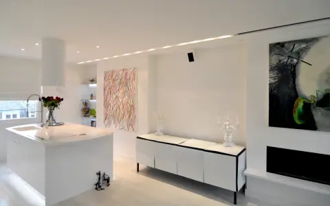 Modernt kök och vardagsrum med vita ytor, konst och spotlights som skapar en ljus och inbjudande atmosfär.