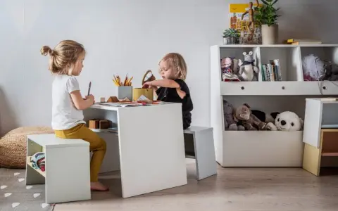 Uroczy kącik dla dziecka w salonie. Dwie dziewczynki rysują przy białym stoliku, otoczone pluszakami i książkami.