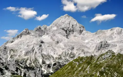 Majestätischer Triglav im Sonnenschein, bereit für die Triglav-Besteigung. Grüne Hänge und Felsen unter blauem Himmel mit Wolken.
