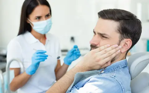 Mężczyzna z bólem policzka u dentysty, który bada jego dziąsła. Może to być objaw problemów z kieszonkami dziąsłowymi.