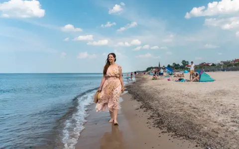 Kobieta w kwiecistej sukience spaceruje brzegiem morza w Sarbinowie. Na plaży widać ludzi odpoczywających na kocach i pod parasolami.