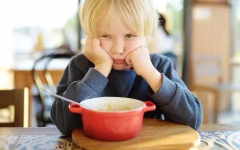 Ein Junge mit blonden Haaren stützt seinen Kopf auf die Hände und schaut lustlos auf eine Schüssel Porridge. Er isst nicht, obwohl er 10 Jahre alt ist.