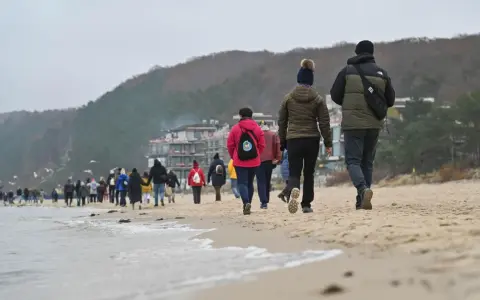 Spacer po plaży w chłodny dzień, idealny na długie weekendy 2026. Grupa ludzi w kurtkach i czapkach idzie wzdłuż morza.