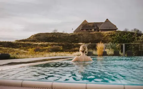 Frau genießt entspannten Wellness Tag in Schleswig-Holstein. Ein idyllischer Pool mit Blick auf Reetdachhaus und Dünenlandschaft.