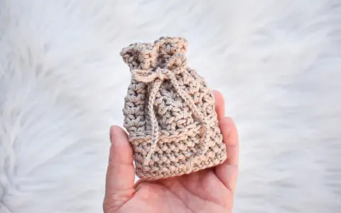 Monedero de ganchillo beige con cordón, sostenido en una mano sobre fondo blanco peludo.
