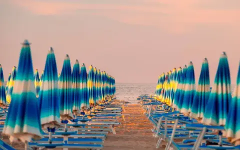 Filetto di ombrelloni a righe blu e bianche sulla spiaggia, con il mare e il cielo rosa all'orizzonte. Un'immagine iconica della riviera adriatica.