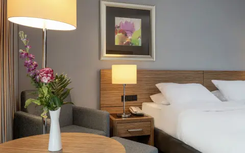Ein Hotelzimmer mit Bett, Nachttischlampe, Sessel und Blumenstrauß.