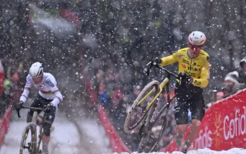 Cyclists battle snowy conditions. Wout van Aert, in yellow, appears to be recovering from a crash, while another rider in white pushes on.