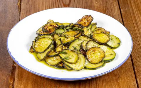 Zucchine alla poverella, fette sottili e dorate, condite con olio e erbe aromatiche, su un piatto bianco con bordo blu.